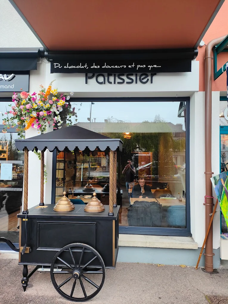 La photo montre un chariot noir au style traditionnel placé devant la vitrine de la boutique. Il possède un petit toit à franges, deux grandes roues en bois et trois cloches en laiton évoquant un stand artisanal. À travers la grande baie vitrée, on distingue l’intérieur chaleureux du magasin et une personne installée à une table. L’ensemble donne une ambiance conviviale et authentique, fidèle à l’esprit de la boutique de Gérardmer.